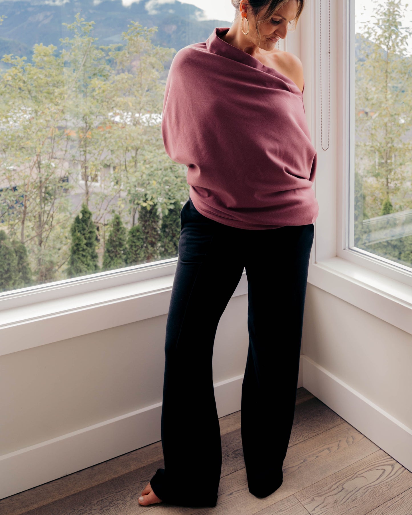 A woman stands barefoot in a bright room corner by large windows, wrapped in a dusty rose shawl or wrap and wearing black pants, with a mountain and tree view outside.