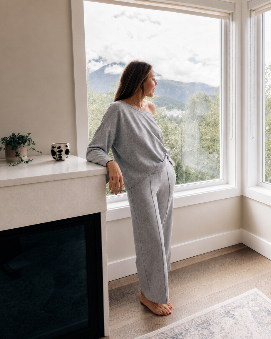 A woman wearing a light gray lounge set stands barefoot by a large window, looking out at a mountain landscape. The set includes a long-sleeve top and matching pants.