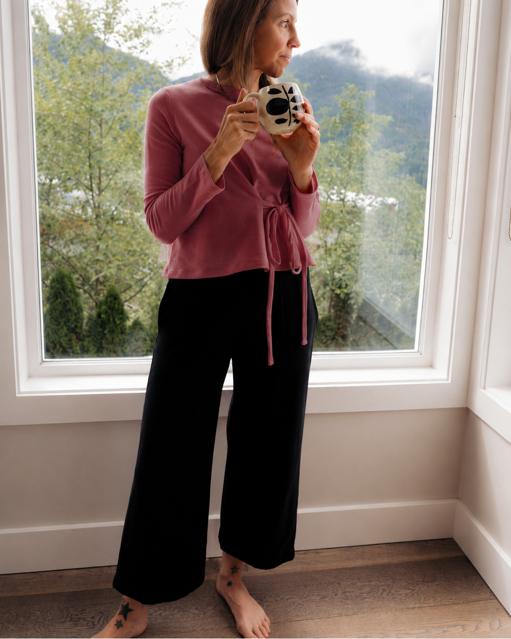 A woman stands barefoot in front of a large window, wearing a dusty rose long-sleeve wrap top and black wide-leg pants, holding a small black and white patterned mug.