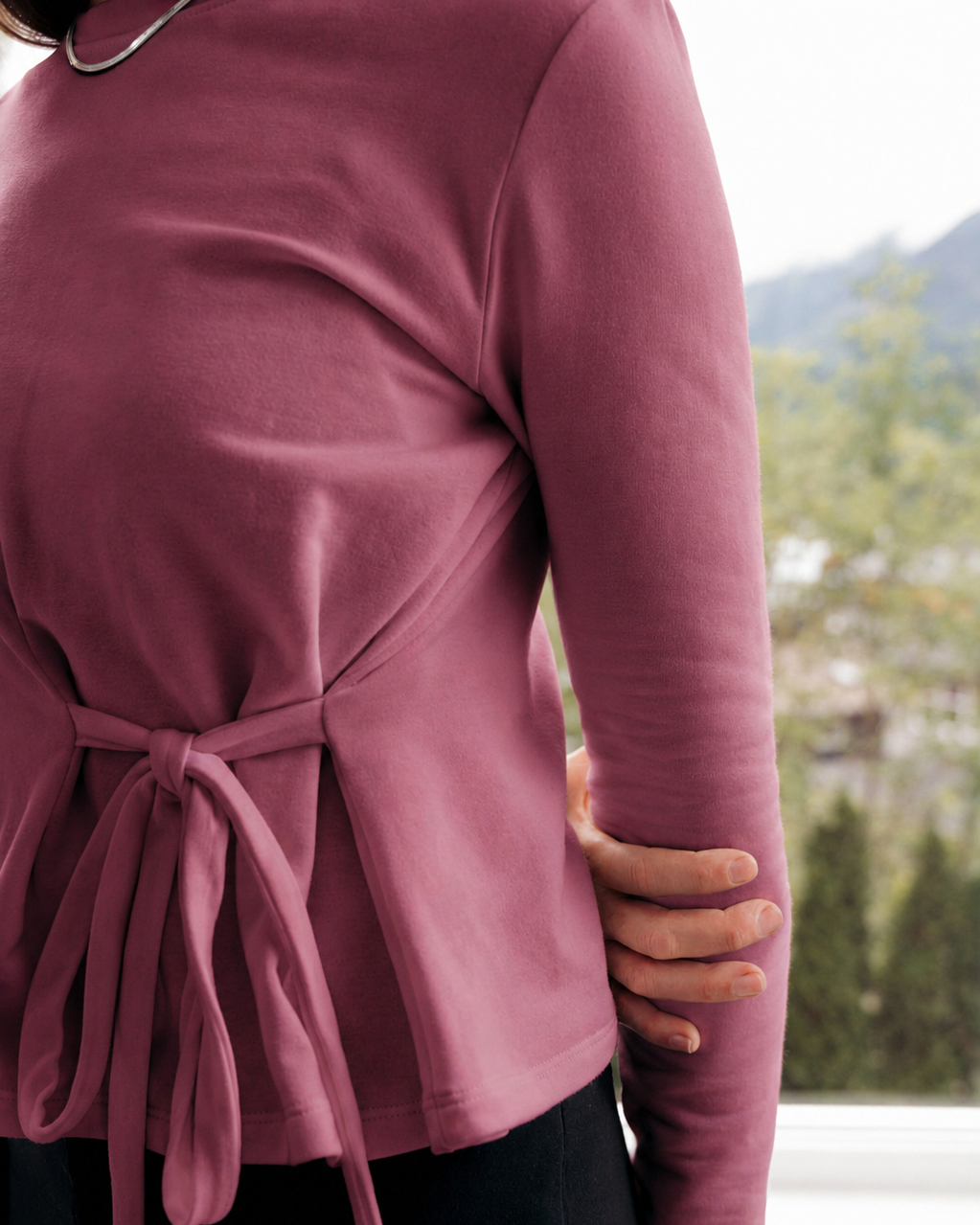 A person wearing a burnt rose long-sleeve wrap-style top, walking with, with a tie waist, paired with black bottoms, Up On, standing near a window with a blurred outdoor background.