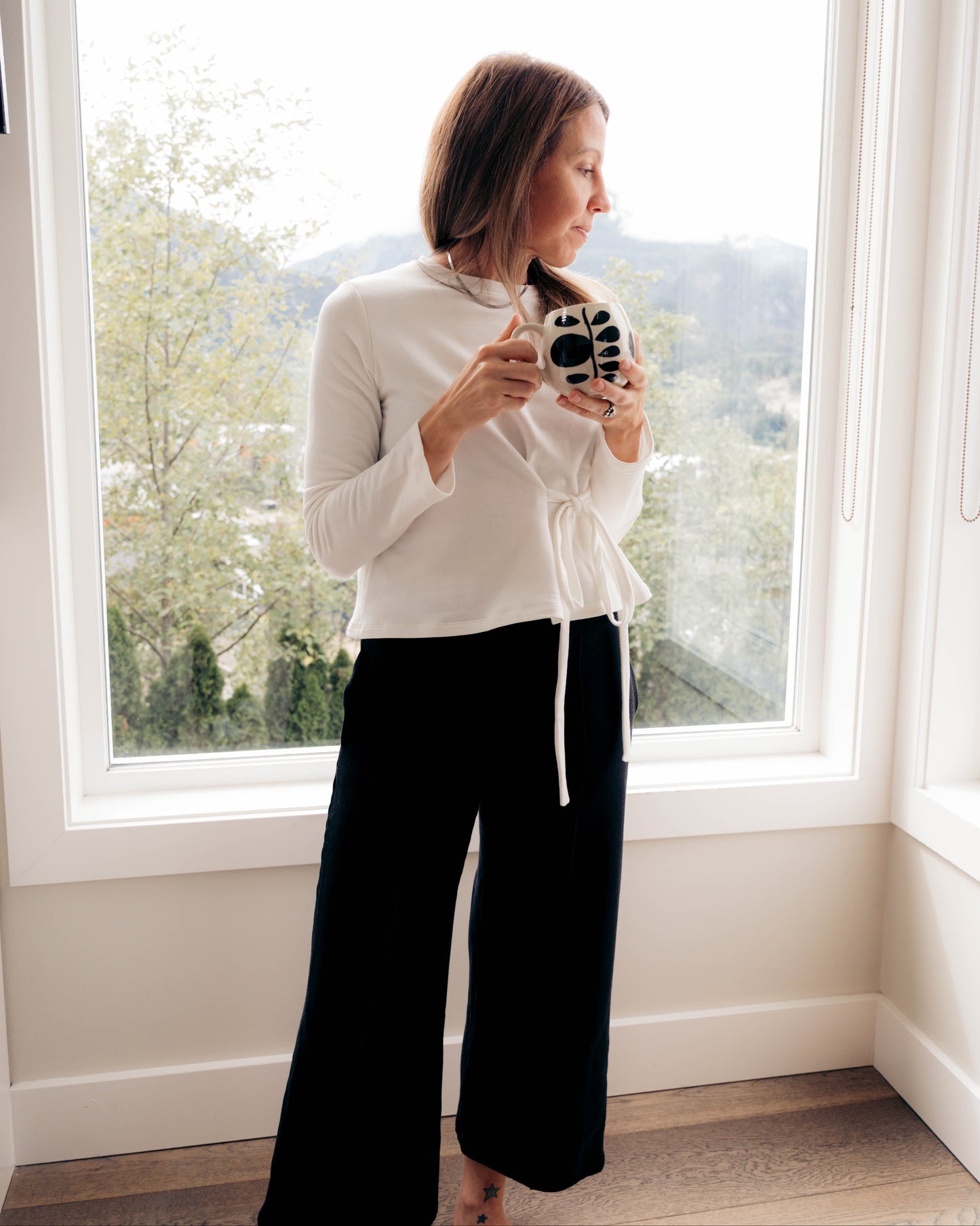 A woman stands in front of a large window wearing an ivory long-sleeve mock neck top, Walking With, with a drawstring and black wide-leg pants, Up On. She is holding a black and white camera.