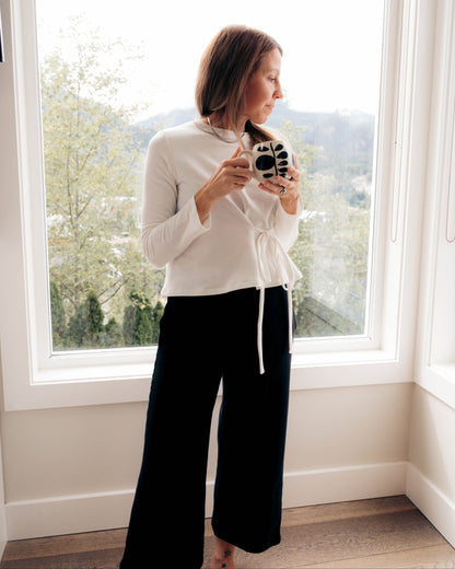 A woman stands in front of a large window wearing an ivory long-sleeve mock neck top, Walking With, with a drawstring and black wide-leg pants, Up On. She is holding a black and white camera.