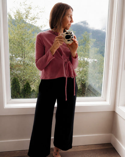 A woman stands barefoot in front of a large window, wearing a dusty rose long-sleeve wrap top called Walking With and black wide-leg soft pants called Up On, holding a small black and white patterned mug.