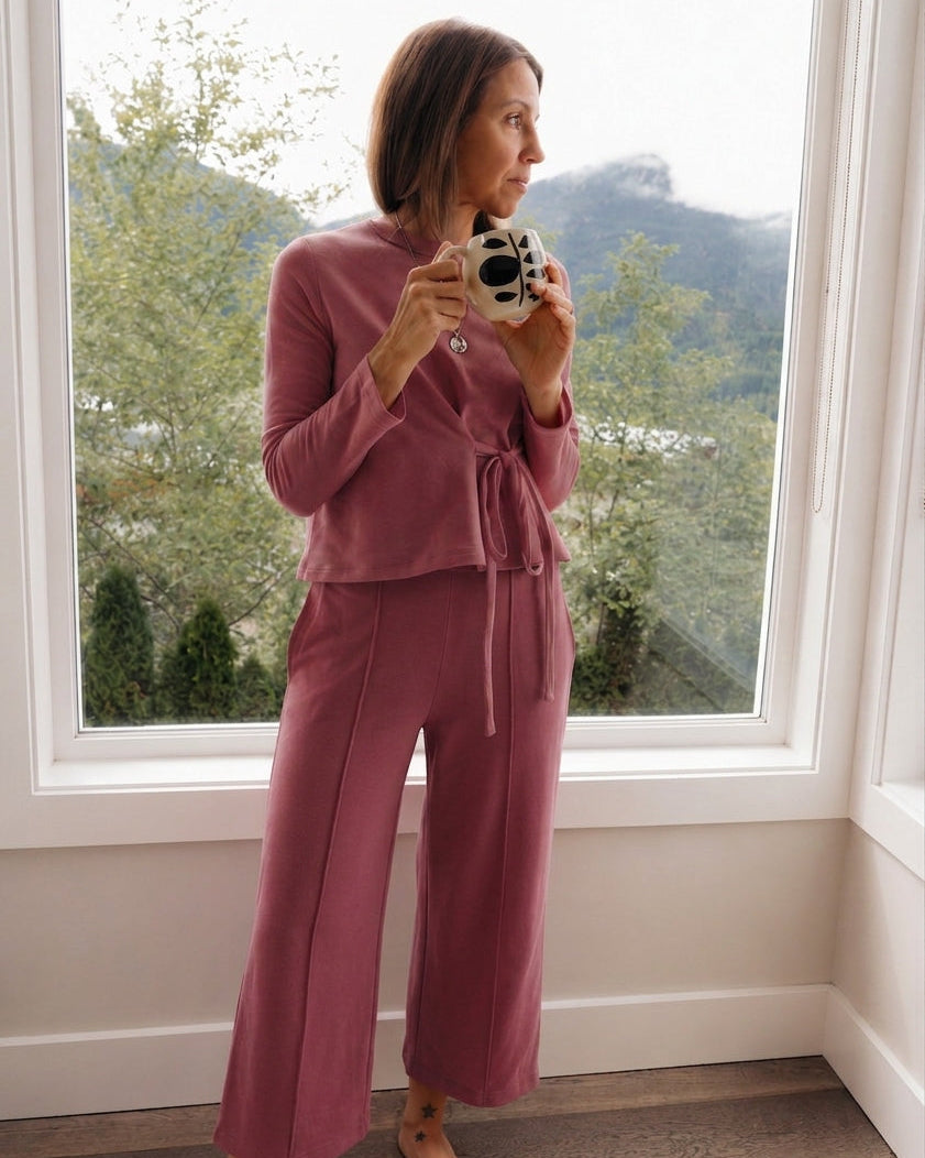 A woman stands barefoot by a large window wearing a dusty rose two-piece lounge set consisting of a long-sleeve cropped top with a tie waist and matching wide-leg pants. She is holding a retro-style camera.