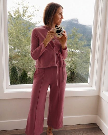 A woman stands barefoot by a large window wearing a dusty rose two-piece lounge set consisting of a long-sleeve cropped top with a tie waist and matching wide-leg pants. She is holding a retro-style camera.