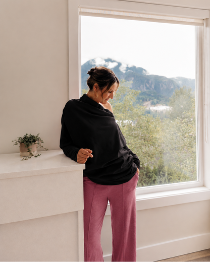 A woman wearing a black cowl-neck top and dusty rose pants stands by a large window with a mountain landscape view, holding a small object in her hand.
