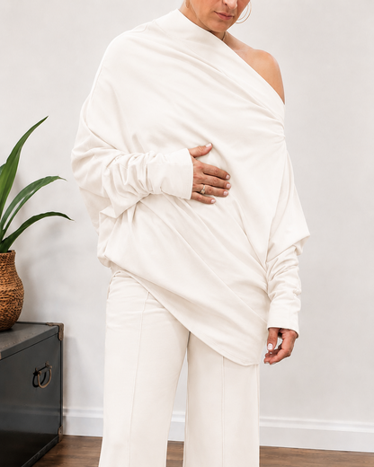 A model wearing a matching off-white two-piece lounge set consisting of a draped long-sleeve top and wide-leg cropped pants, standing on a wooden floor.