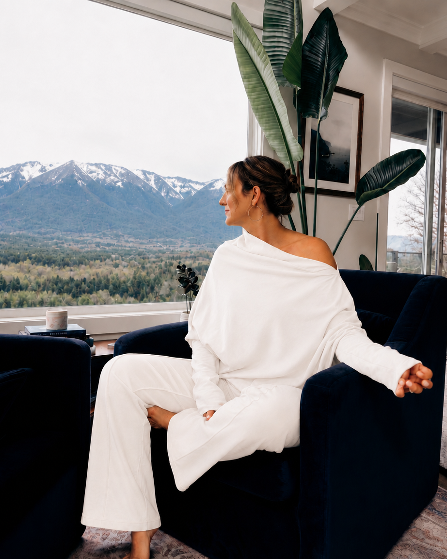 A woman sits in a dark blue armchair by a large window, wearing a white one-shoulder top and matching wide-leg pants. Outside the window, a mountain landscape with snow on the peaks is visible.