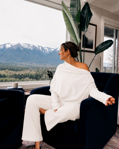 A woman sits in a dark blue armchair by a large window, wearing a white one-shoulder top and matching wide-leg pants. Outside the window, a mountain landscape with snow on the peaks is visible.
