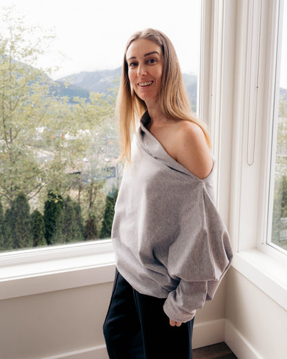 A woman with blonde hair stands by a large window wearing a light gray knit sweater draped off one shoulder and black pants. The background shows a bright outdoor view with trees and hills.