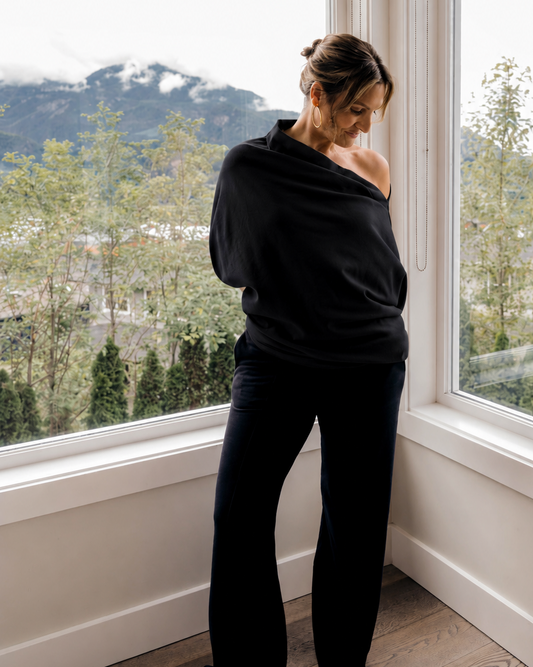 A woman stands barefoot in a bright room by large windows, wearing a black off-the-shoulder top and black wide-leg pants, with a mountain and tree landscape visible outside.