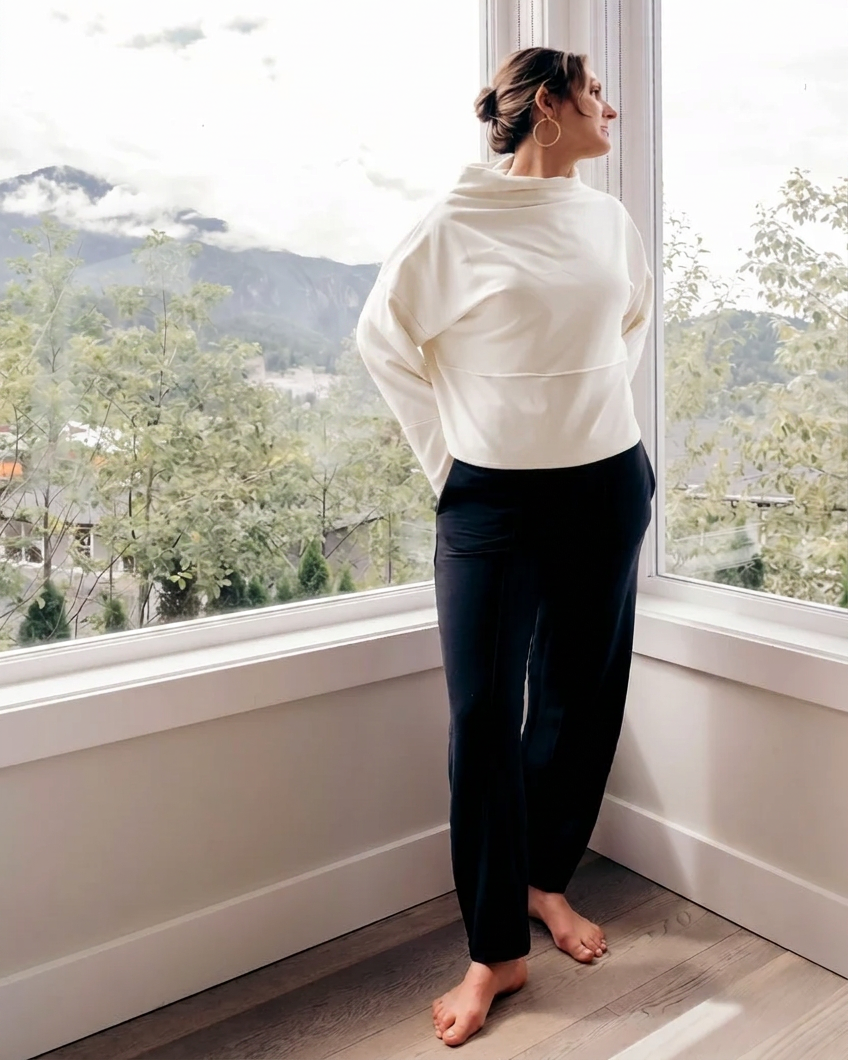 A woman stands barefoot by a large window, wearing a cream-colored long-sleeve cropped hoodie and dark navy pants, looking out at a scenic mountain landscape.