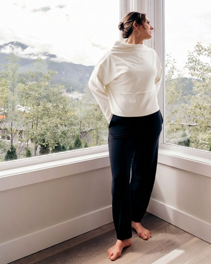 A woman stands barefoot by a large window, wearing a cream-colored long-sleeve cropped hoodie and dark navy pants, looking out at a scenic mountain landscape.