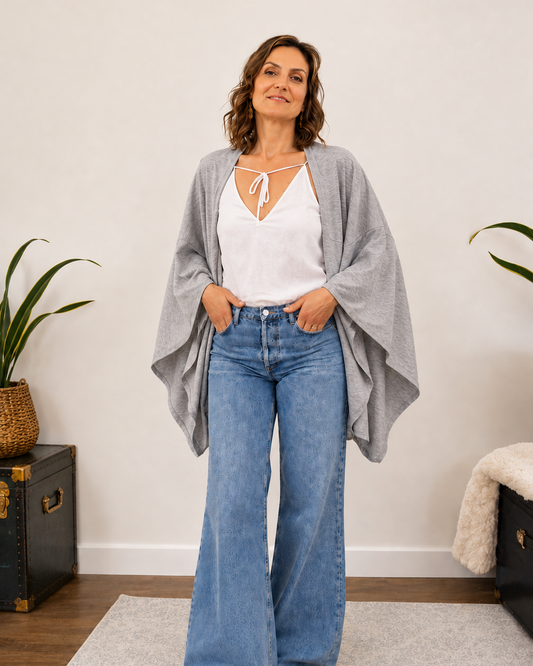 A woman stands indoors wearing a light gray open-front poncho-style cardigan over a white tie-front top and wide-leg blue jeans, with tan sandals. The room has a white wall, wood floor, a rug, and potted plants.