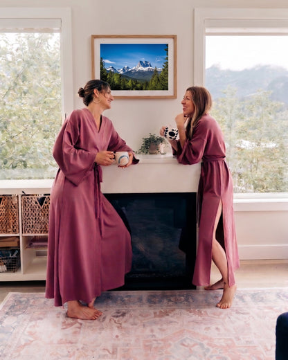 Two women wearing matching mauve-purple long robes stand barefoot by a fireplace in a bright room, holding mugs and smiling at each other. A framed mountain landscape print hangs above the fireplace, with large windows and trees visible outside.