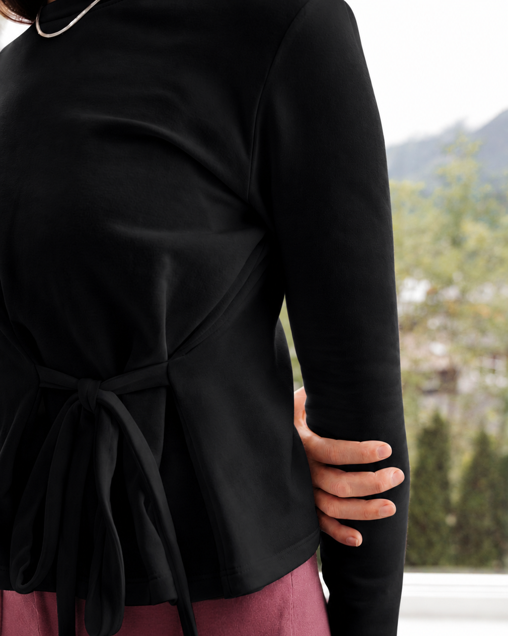 A close-up of a person wearing a black long-sleeve wrap-style top with a self-tie waist belt, paired with dusty rose pants. The background shows a softly blurred outdoor window view.