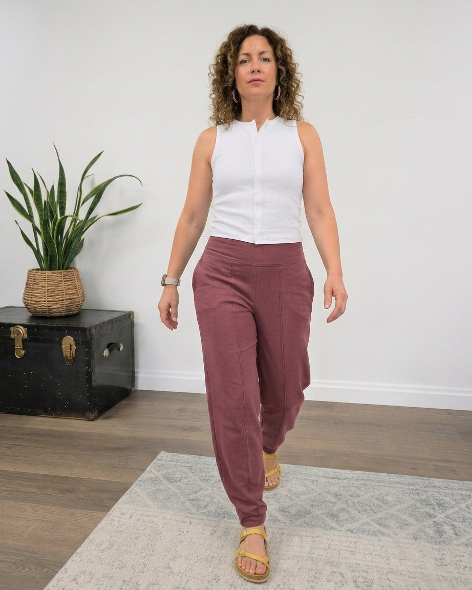 A woman walking indoors wearing a sleeveless white top and dusty rose pants, standing on a light patterned rug with a plant and black trunk against a white wall.